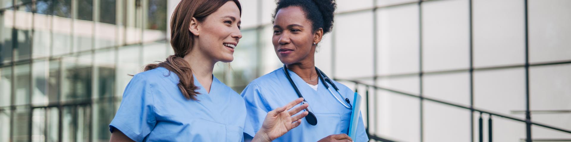 Two happy female doctors walking in front of hospital building and communicating