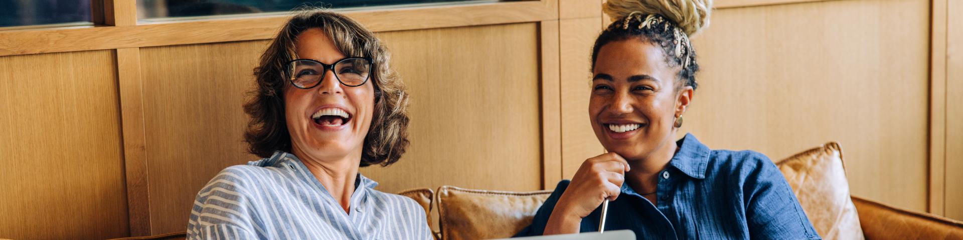 Two women sit together on a leather couch, smiling and engaging while working on a laptop. The setting exudes warmth, teamwork, and positivity, creating an inviting and productive atmosphere.