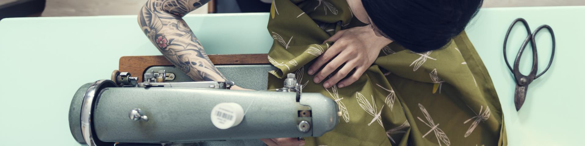 Overhead view of a female dressmaker working on a sewing machine in her design studio. Colour, horizontal format with a very shallow focus point on her hands as she works feeding the material through the machine. She is wearing a white sleeveless top that shows of her full sleeve tattoo she is half Japanese half european with dark hair.