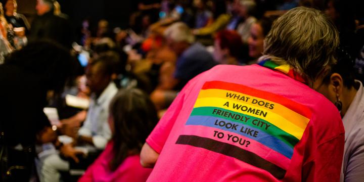 Person trying to find a seat in a crowded theatre wearing a shirt saying "what does a women friendly city look like to you?"