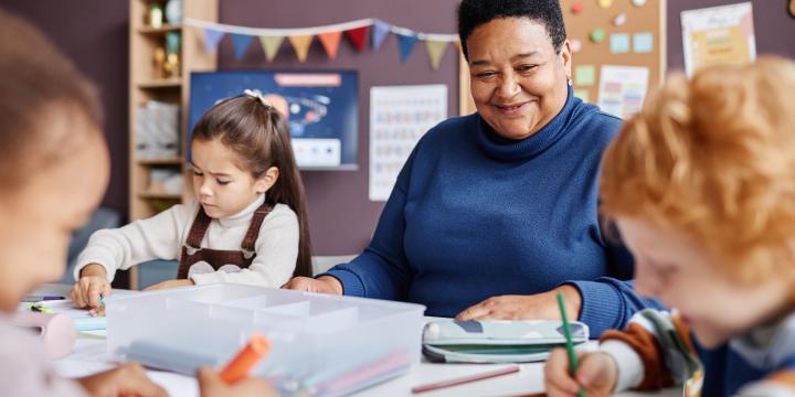 Kindergarten teacher seating on a kids table while kids draw with crayons