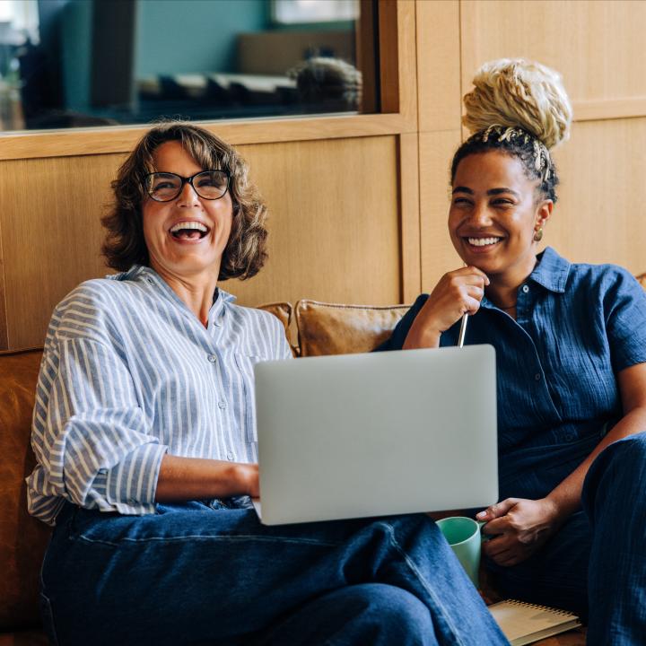 Two women sit together on a leather couch, smiling and engaging while working on a laptop. The setting exudes warmth, teamwork, and positivity, creating an inviting and productive atmosphere.