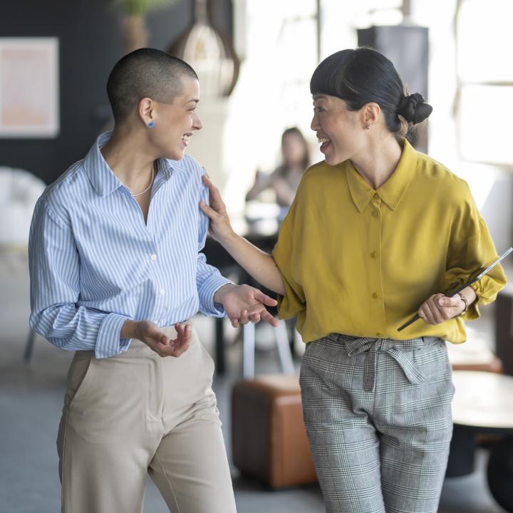 Two businesswomen are walking and talking together in a modern office, holding a tablet and smiling